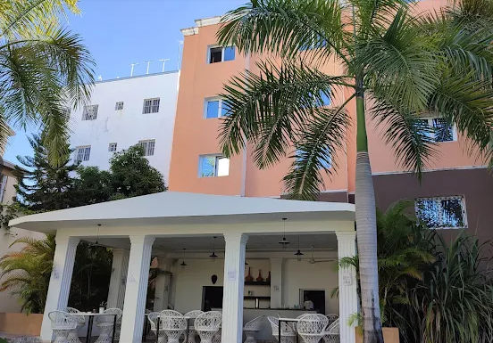 Vista exterior de una terraza sombreada con mesas y sillas de ratán blanco, frente a un edificio hotelero de varios niveles en tonos durazno, blanco y café, rodeado de vegetación tropical en Punta Cana.