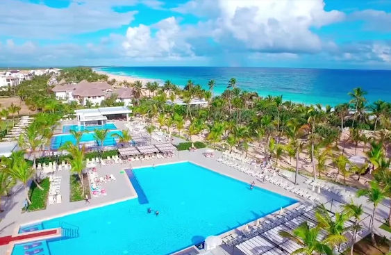"Vista aérea de un complejo turístico de lujo frente al mar con grandes piscinas, palmeras y playa de arena blanca bajo un cielo azul"