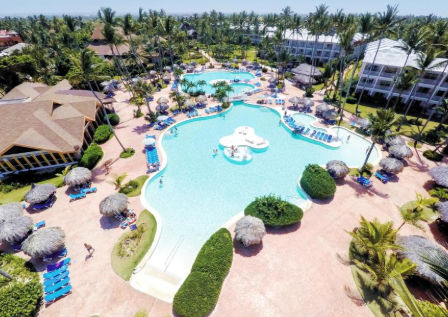 Aerial view of a tropical resort with a large, free-form swimming pool, palm trees, sun loungers, and traditional beach umbrellas.