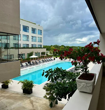 "Vista desde balcón de hotel en Punta Cana con planta de flores rojas en primer plano, piscina rectangular de agua cristalina rodeada de camastros y moderno edificio del complejo hotelero al fondo bajo un cielo nublado."