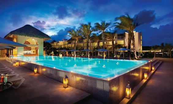 "Night view of a large infinity pool at a luxury tropical resort, illuminated with blue lights and surrounded by lit decorative lanterns. In the background, a modern hotel building can be seen next to a thatched-roof structure (palapa), palm trees, and a 