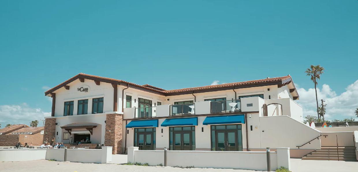 La Casa Del Mar seen from the sand, a two-storey beachfront building with blue awnings and an open terrace.
