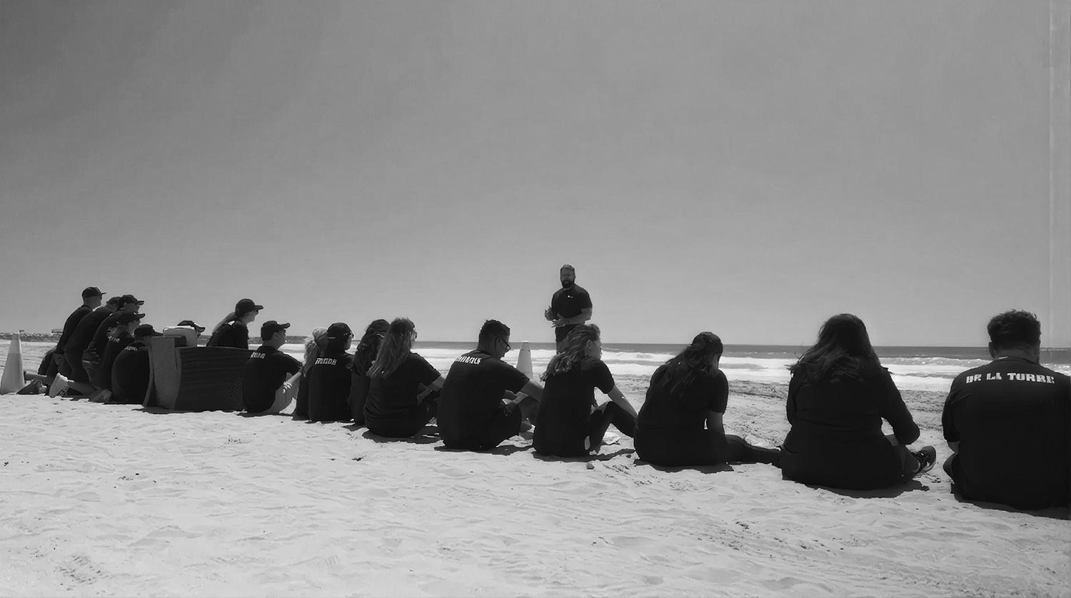 A group sitting on the sand in a long line, listening to an instructor with the Pacific behind him.