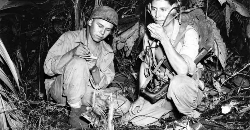 Two Marines crouched in jungle undergrowth, one writing, one working a field radio.