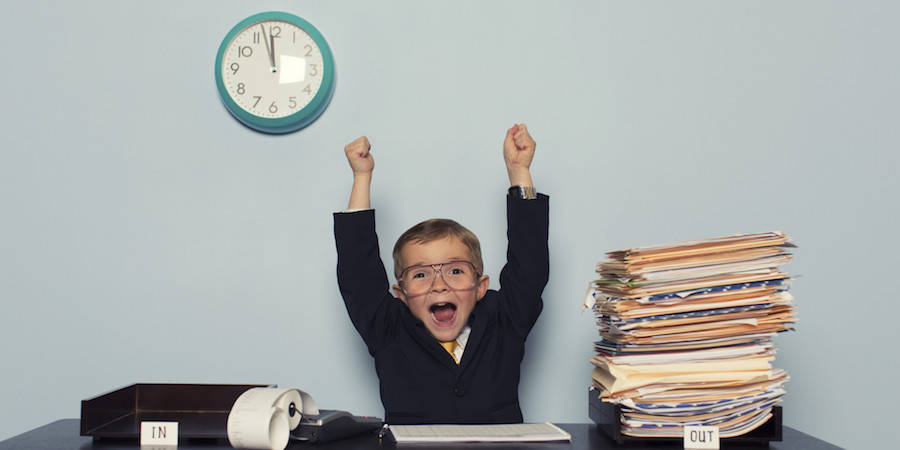 little boy sitting at a work desk in a business suit wearing glasses and a huge stack of papers looking excited