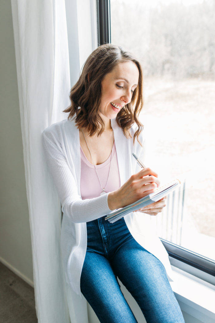Janelle Reichman of Ellanyze sitting on the window sill in her office writing in a notebook with pencil