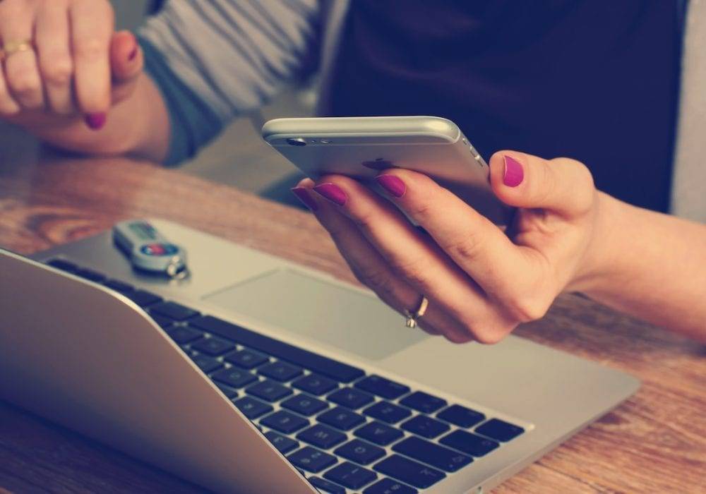 make website mobile friendly: woman sitting at her laptop with her phone in the hand