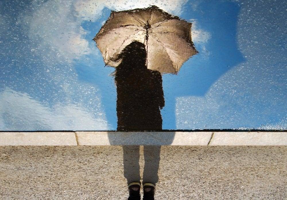 life before COVID: reflection in a puddle of a woman holing an umbrella, bright blue sky behind her