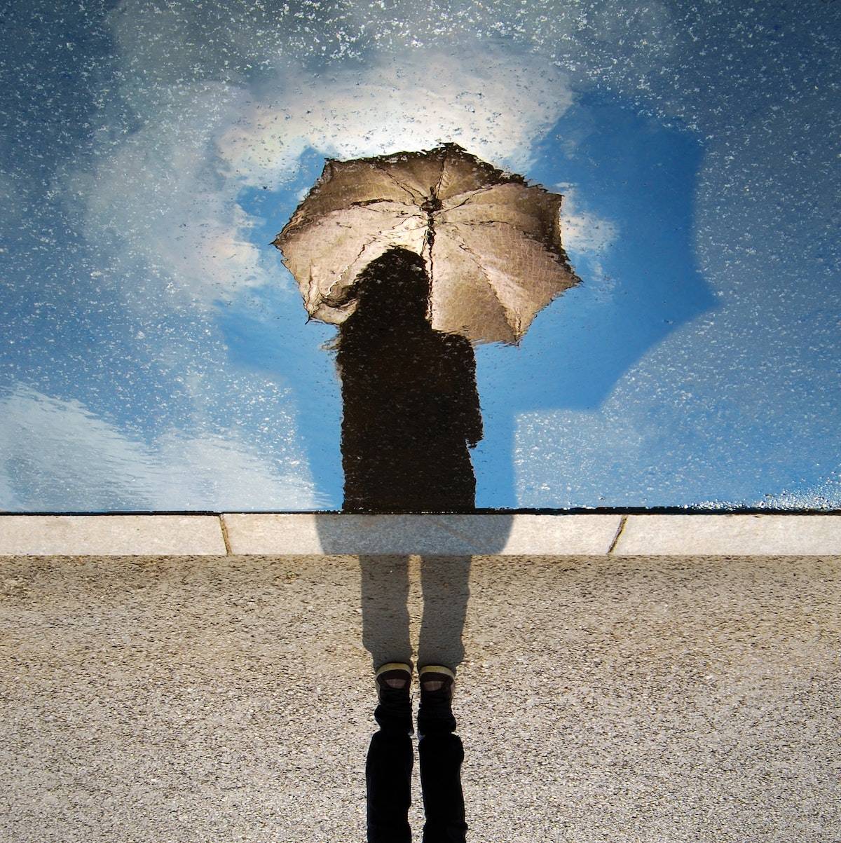 life before COVID: reflection in a puddle of a woman holing an umbrella, bright blue sky behind her