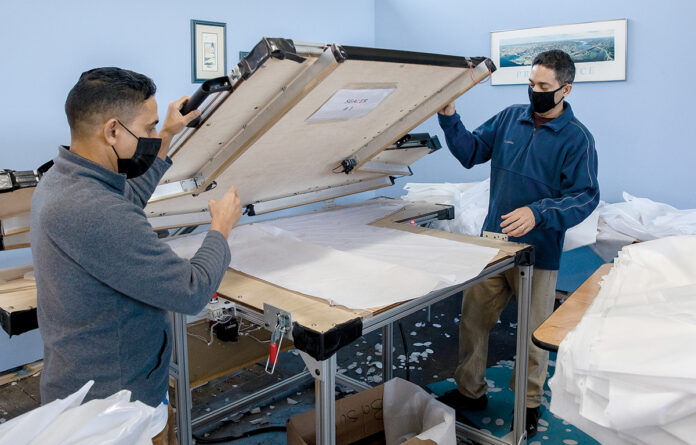 SWITCHING GEARS: Felis Franco, left, and Juan Arrojo load a sealer at Eco Global Manufacturing LLC in Providence. During the COVID-19 pandemic, the product design company switched gears to produce face shields and isolation gowns. / PBN PHOTO/TRACY JENKINS SWITCHING GEARS: Felis Franco, left, and Juan Arrojo load a sealer at Eco Global Manufacturing LLC in Providence. During the COVID-19 pandemic, the product design company switched gears to produce face shields and isolation gowns. / PBN PHOTO/TRACY JENKINS
