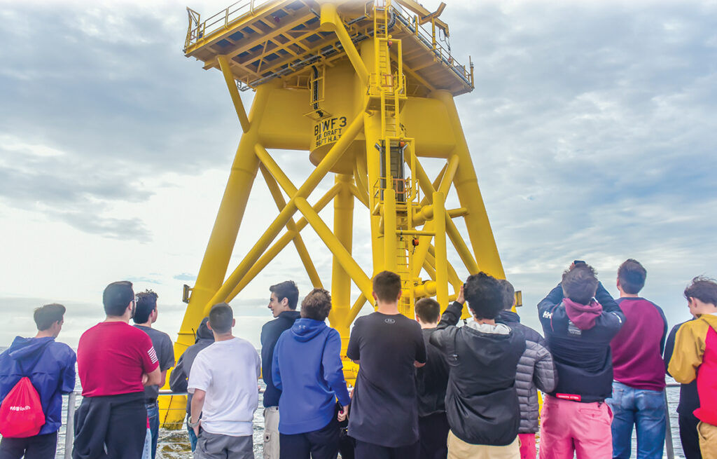 LOOMING LARGE: A boat carrying a group of Rhode Island high schoolers circles the base of one of the massive turbines of the Block Island Wind Farm. Students have been participating in Wind Win RI, a career pathway program for the offshore wind industry. / COURTESY DOUG LEARNED/LUCKY DAWG PHOTOGRAPHY