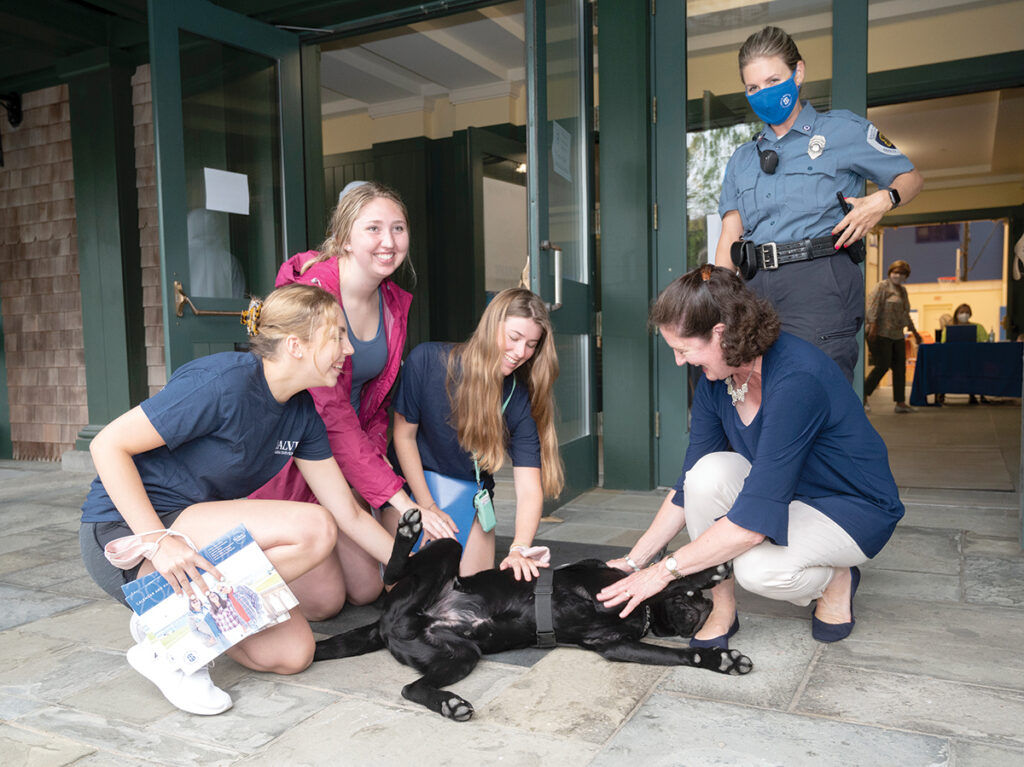 NEW ADDITION: Salve Regina University President Kelli J. Armstrong joins students Katie Mountcastle, Julia DelMastro and Julie Burgjohann, all juniors, to pet Salve’s new community resource dog, Ruggles, a 10-month-old black Lab. In the background is Campus Safety and Security officer Michelle Caron. / PBN PHOTO/DAVE HANSEN