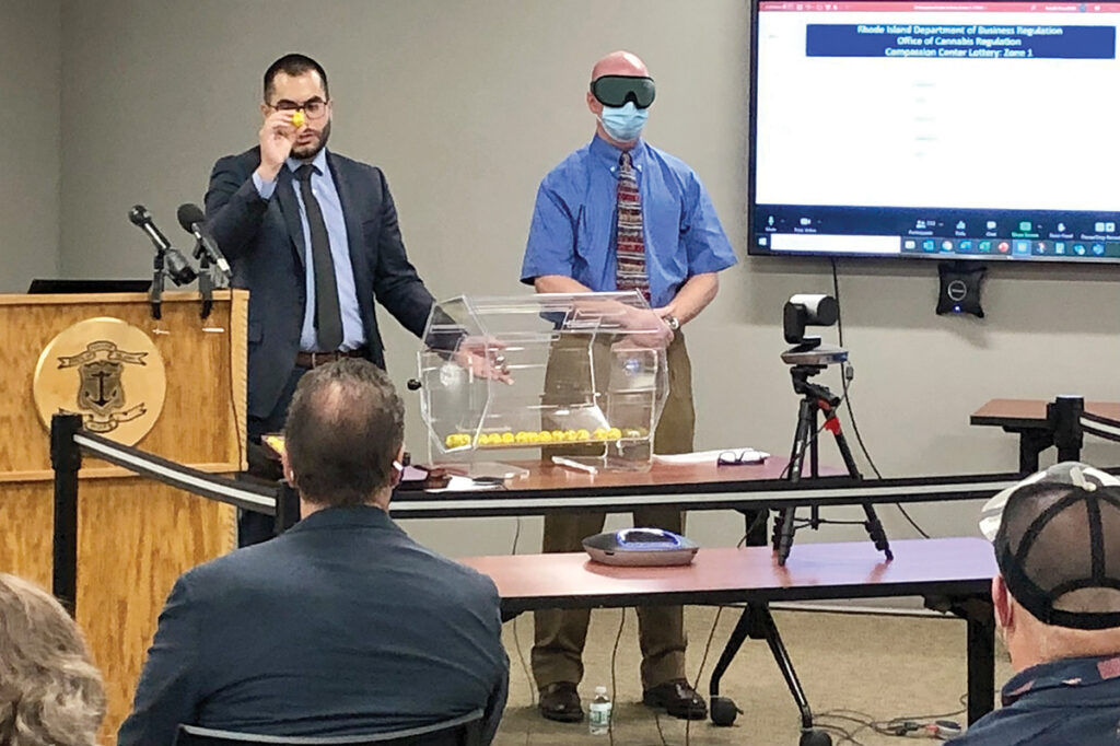 A MATTER OF LUCK: Matthew Santacroce, left, the state’s cannabis chief, displays the first winning ball during a lottery to award five licenses for medical marijuana dispensaries on Oct. 29. At right is Russ Griffiths, an R.I. Department of Business Regulation official who picked the balls from a tumbler while blindfolded. / PBN FILE PHOTO/CASSIUS SHUMAN