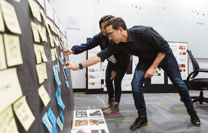 STEADY GROWTH: Richard Davia, managing director of creative and branding at (add)ventures, and Carli Kabilyo, assistant director of design and branding, add Post-it notes to a board at the company’s new offices in East Providence. / PBN PHOTO/RUPERT WHITELEY