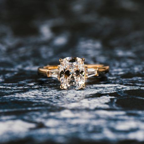 Main Keyword how much should you spend on an engagement ring - image of old mine brilliant cut elongated cushion lab grown diamond engagement ring with baguette side stones on abstract blue black white background