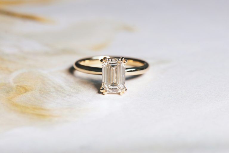 how much should you spend on your engagement ring: lab grown diamond emerald engagement ring with double raindrop prongs and 14k yellow gold band. Abstract hint of yellow background.