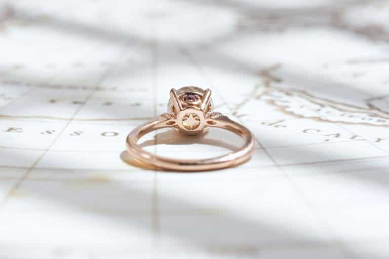 A round-cut rose gold engagement ring featuring a compass logo under the center stone. A tiny rhodolite garnet and emerald are on each side, placed atop a crescent.