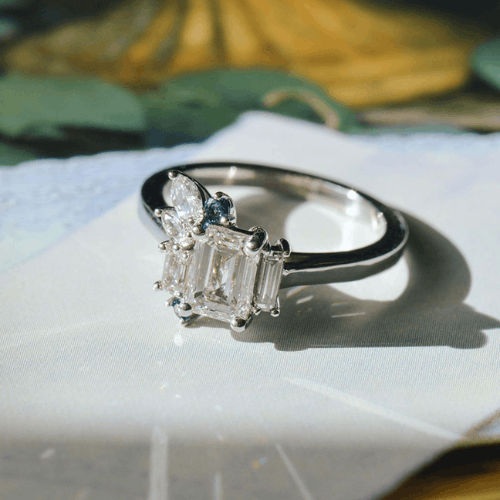 A lab-grown emerald-cut diamond ring with two straight baguette cuts (one longer than the other). Atop the shorter baguette sits a pear and marquis diamond, and a round blue sapphire.