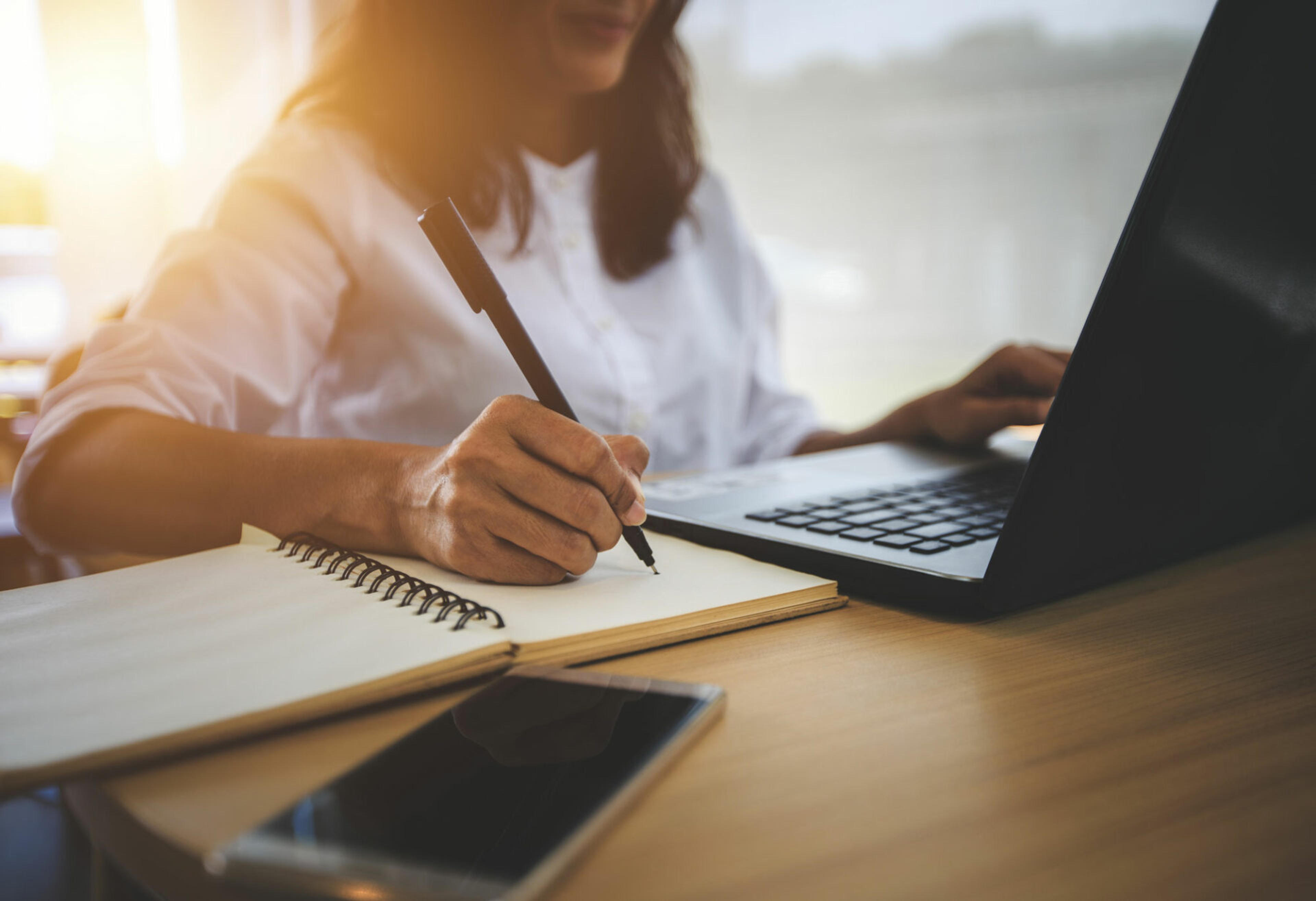Young woman sitting at a desk and taking notes while reading from her laptop.