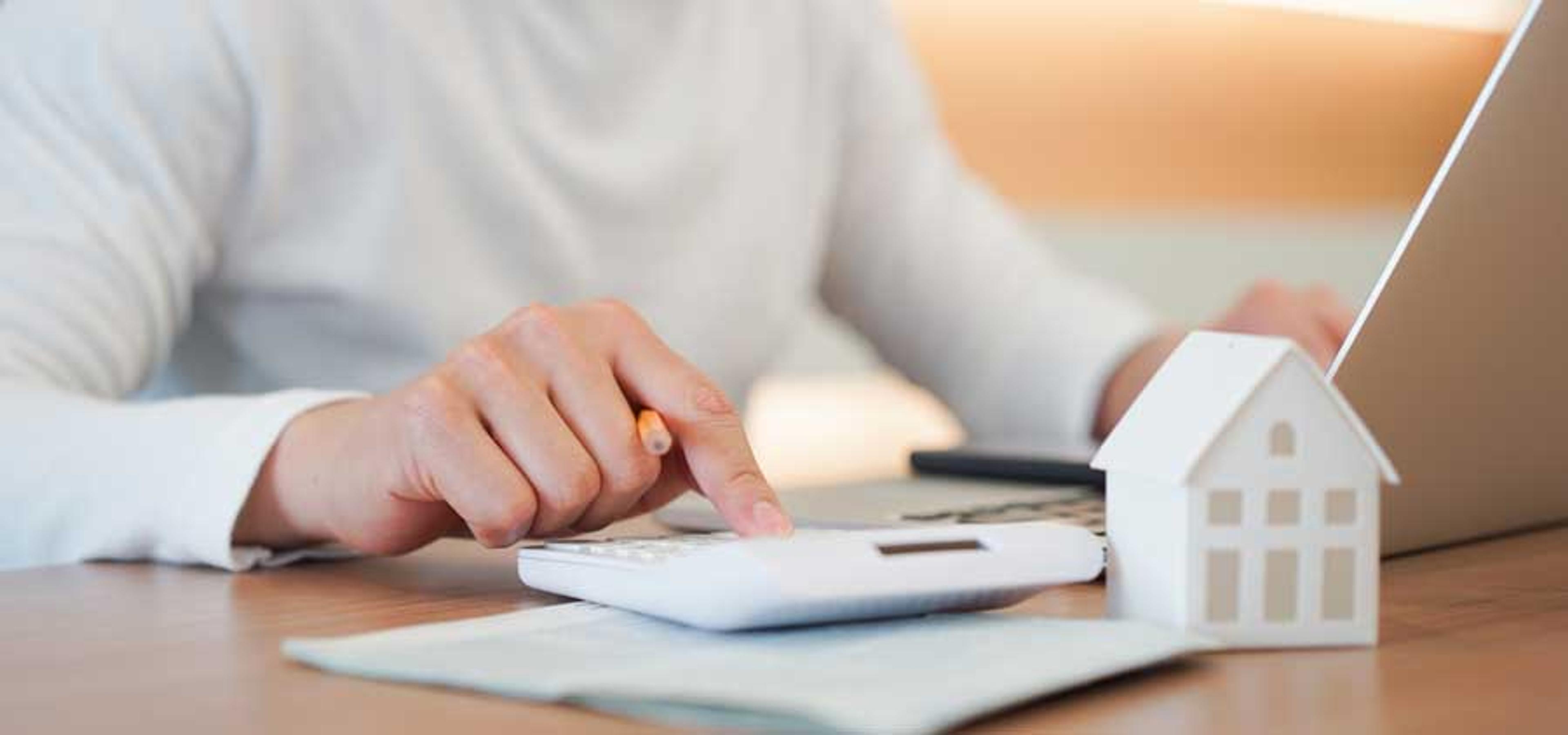 A person with a pencil, notepad, and calculator in front of a computer and miniature house model