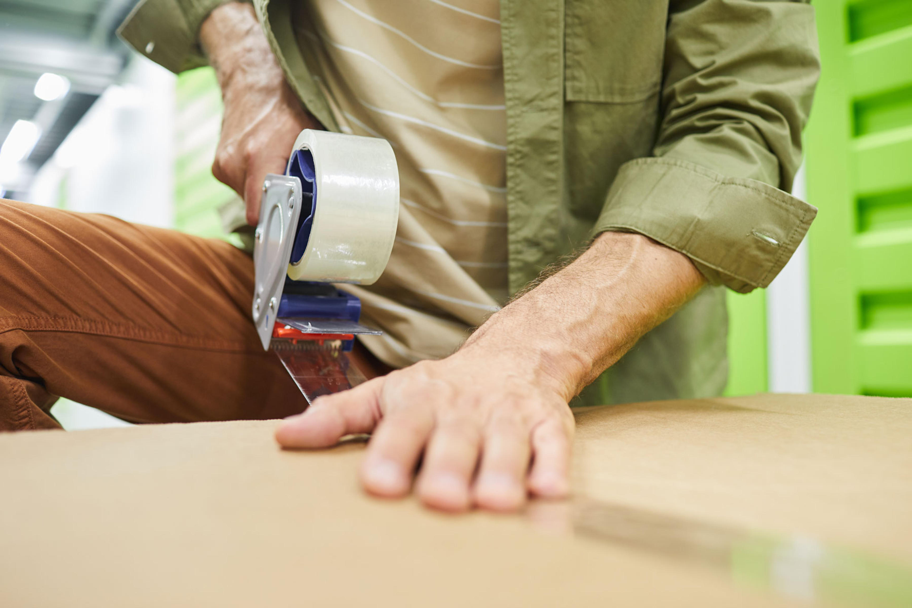 Close up of unrecognizable man sealing cardboard boxes with scotch tape gun while packing in self storage unit