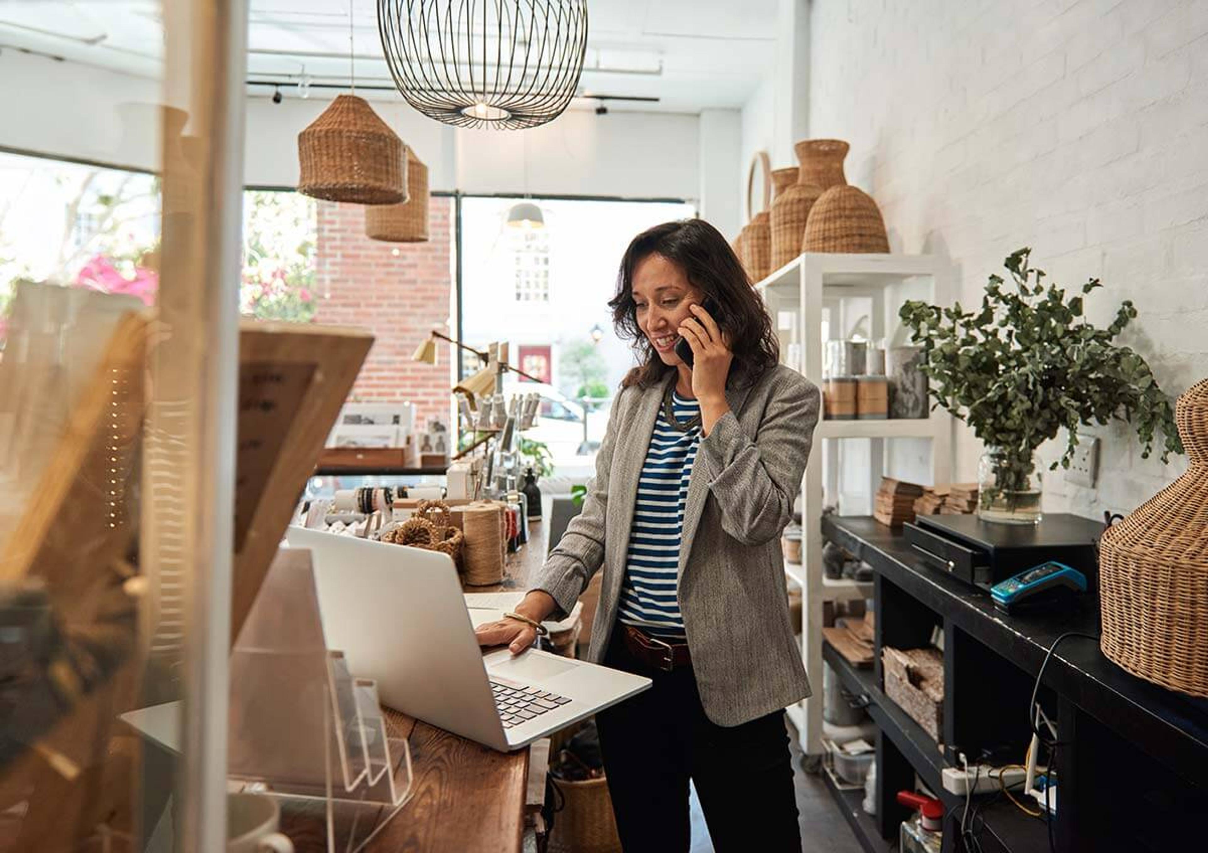 Female shop owner standing behind counter while speaking on the phone and looking at her laptop computer