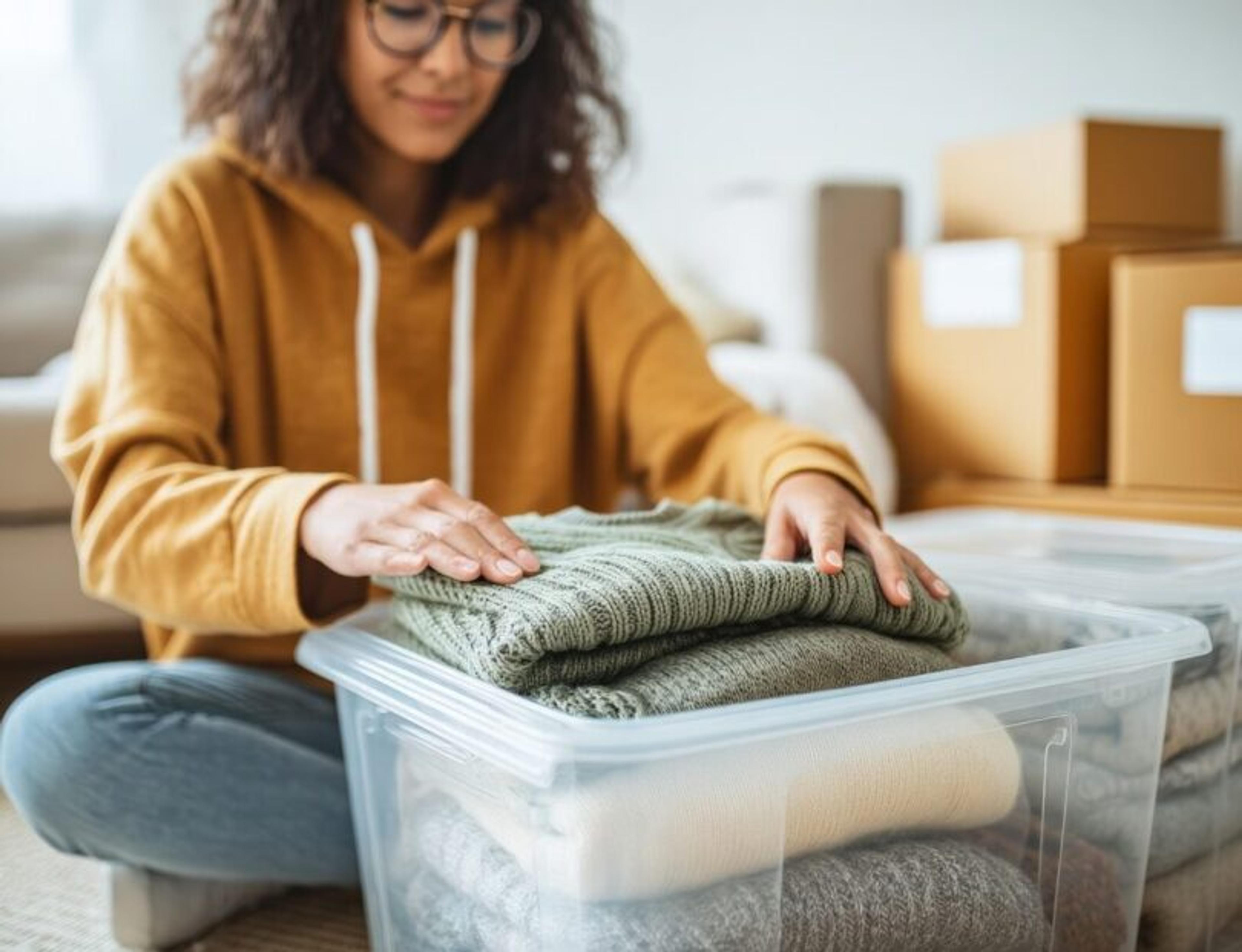 Woman sitting on the floor folding sweaters into a clear plastic storage bin, with packed moving boxes in the background.