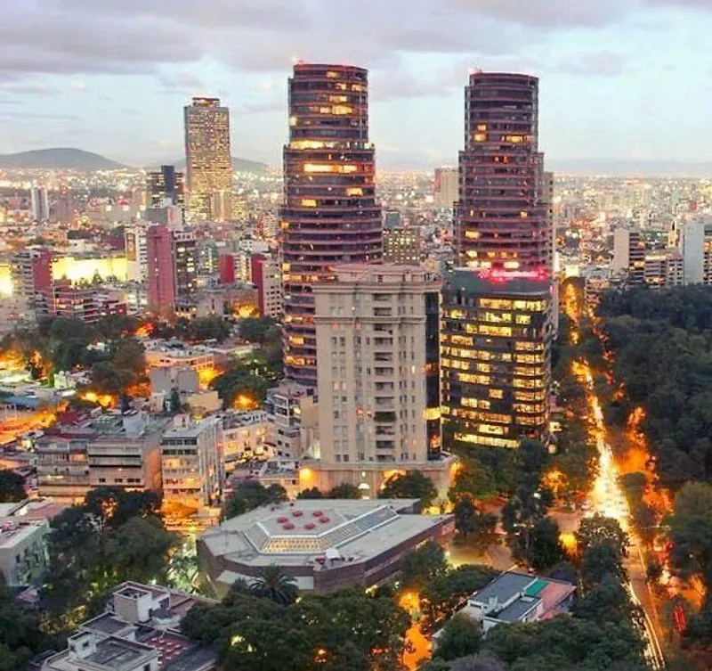 Night cityscape with three illuminated tall buildings in the center, surrounded by smaller structures and trees