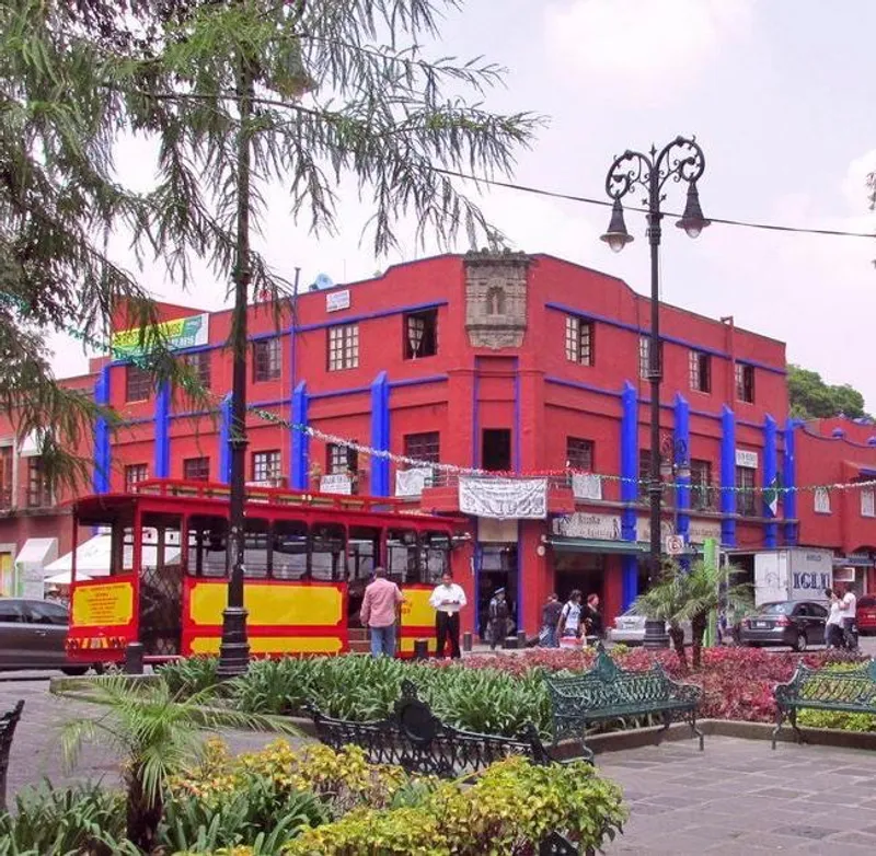 A red building with blue trim in Coyoacán, featuring a red and yellow trolley, benches, and flowers
