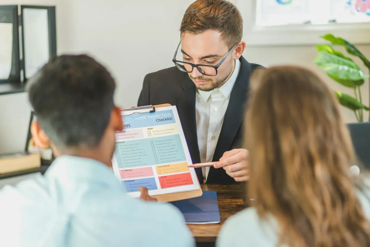 A man in a suit and glasses reviews a colorful inspection checklist on a clipboard with two other people at a desk