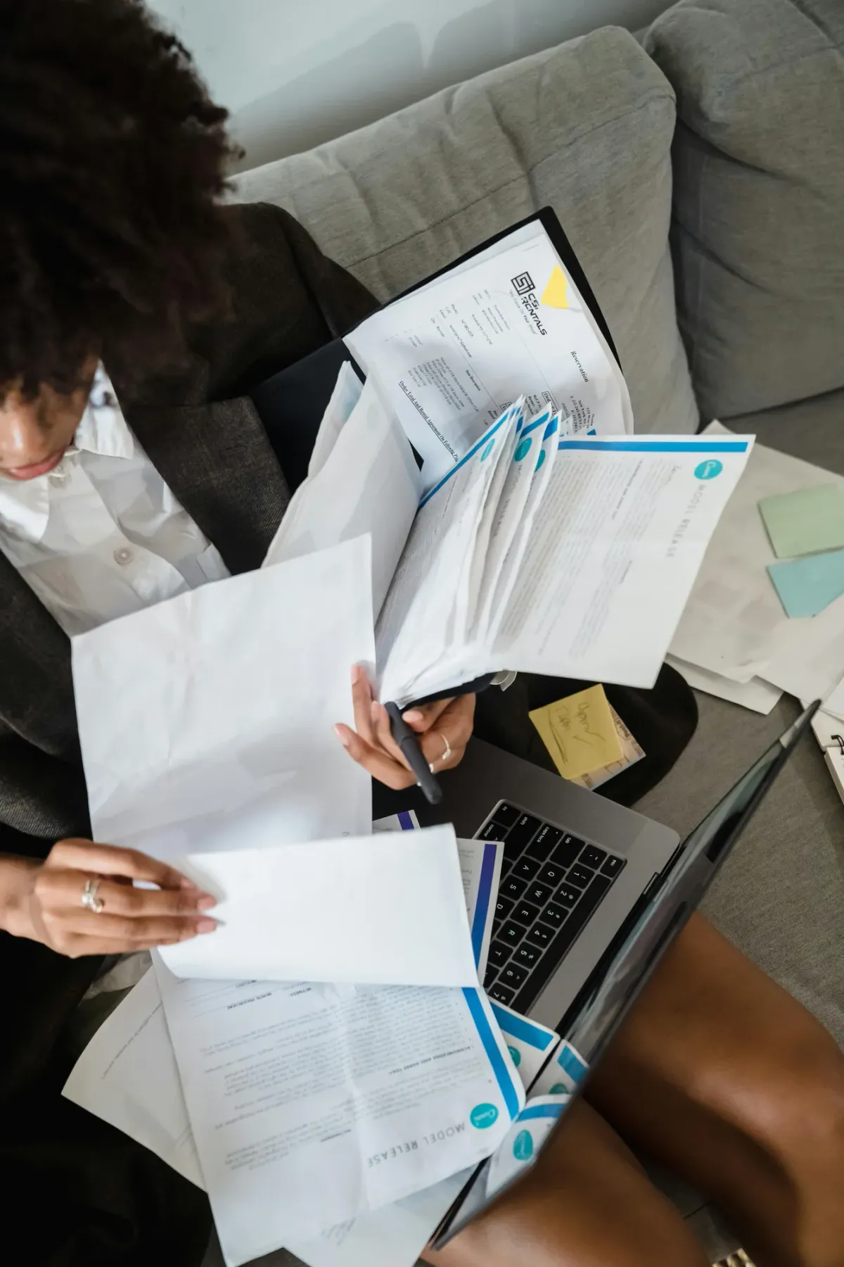 An overhead view of a person in a blazer reviewing stacks of paperwork and invoices beside an open laptop