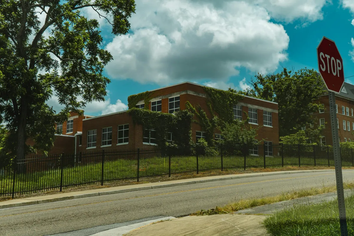 A two-story brick school building partly covered in ivy, seen from across the street behind a black metal fence with a stop sign
