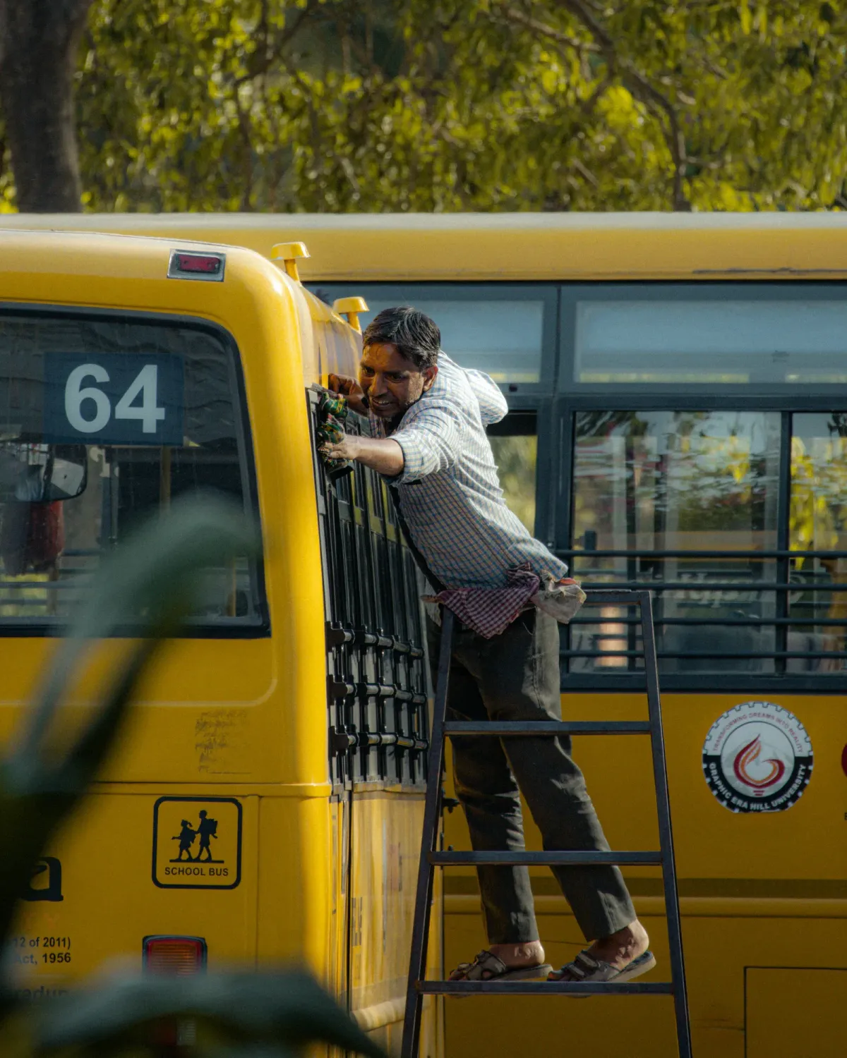 A worker on a step ladder repairs the exterior of a yellow school bus numbered 64