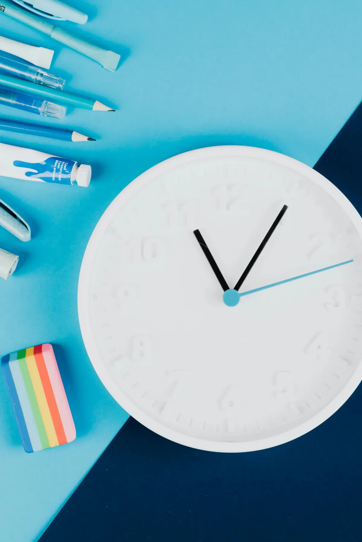 A white wall clock beside a flat-lay of pens, pencils, paint tubes, and a rainbow eraser on a two-tone blue background