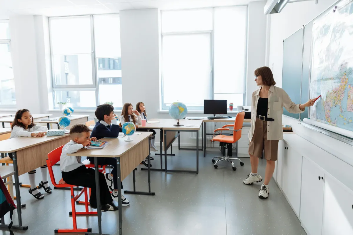 A teacher standing at a wall map points toward young students seated at desks in a bright classroom with globes on the windowsill