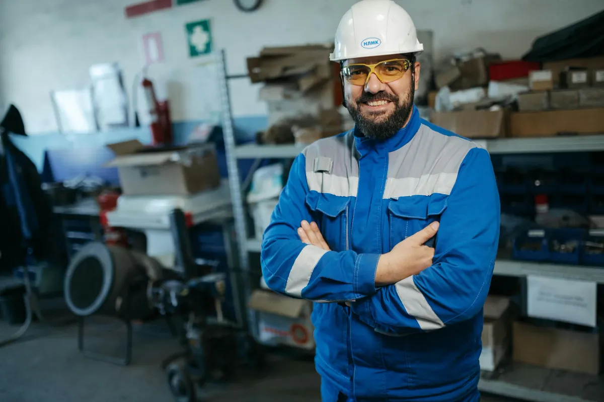A facilities worker in a white hard hat and blue uniform standing with arms crossed in a storage area