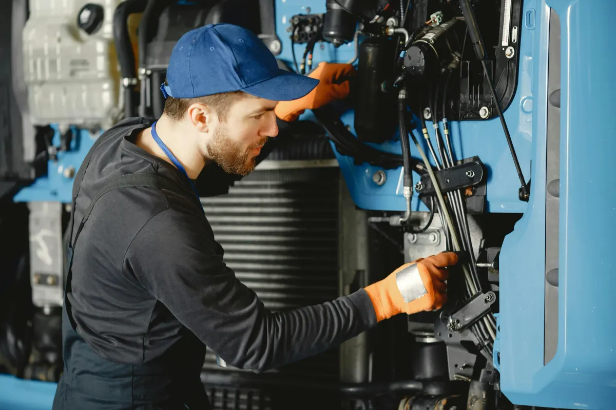 A maintenance technician in a cap and work gloves inspecting mechanical equipment with a flashlight