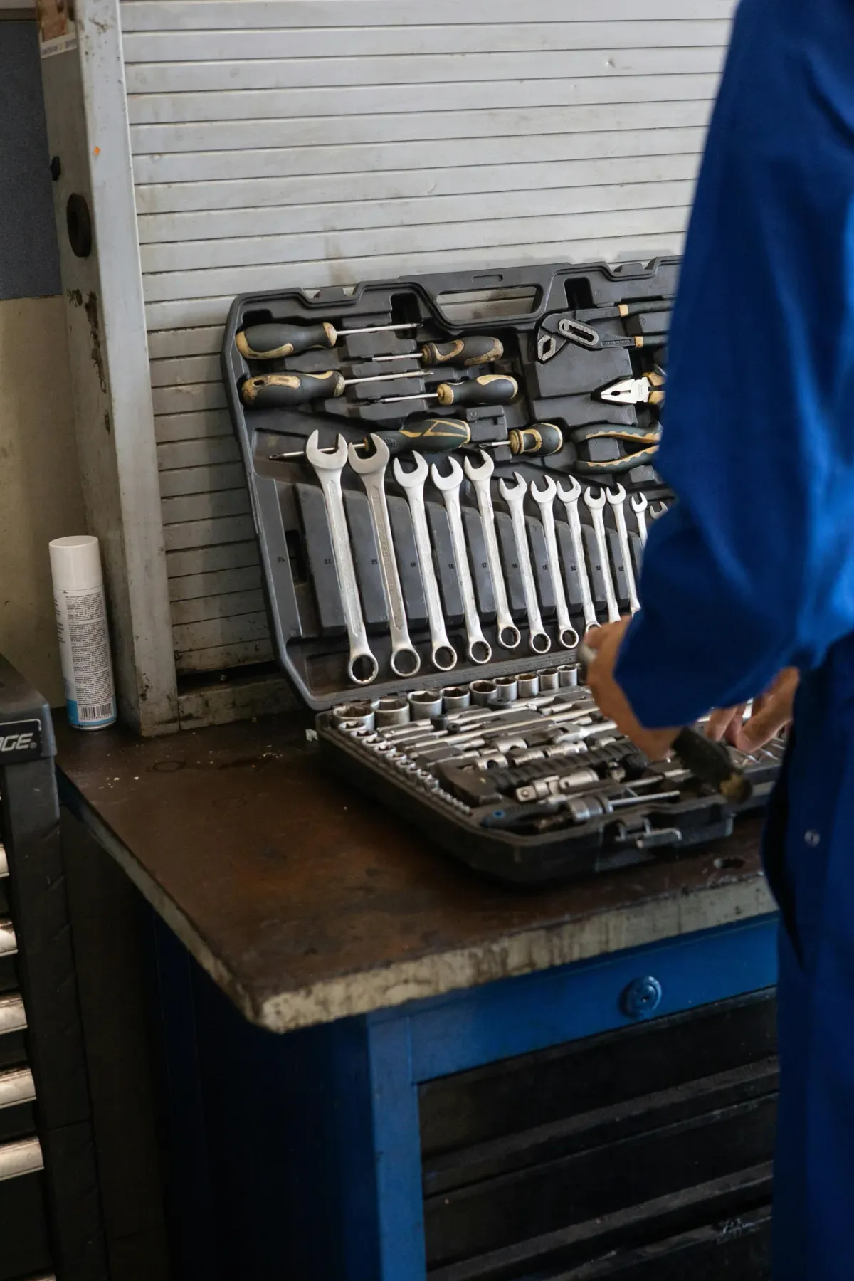 A worker in a blue uniform carrying a tray of metal parts beside a workbench