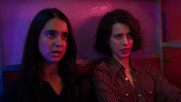 Margaret Qualley and Garaldine Viswanathan sitting beside each other in a bar in Drive-Away Dolls