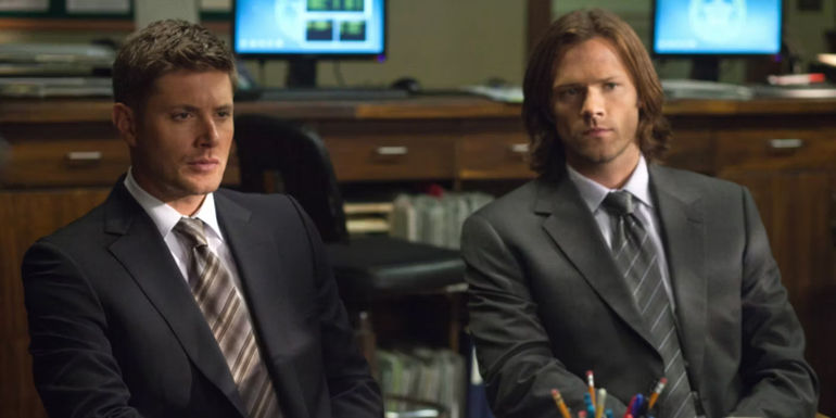 Dean and Sam in suits sitting behind a desk in Supernatural season 8