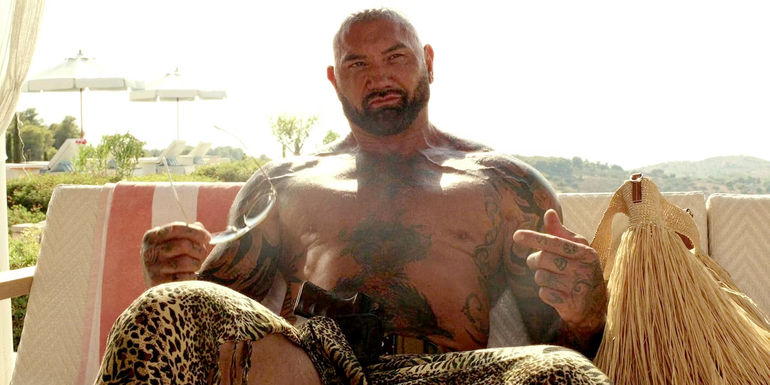 Dave Bautista sitting on a patio shirtless in Glass Onion A Knives Out Mystery