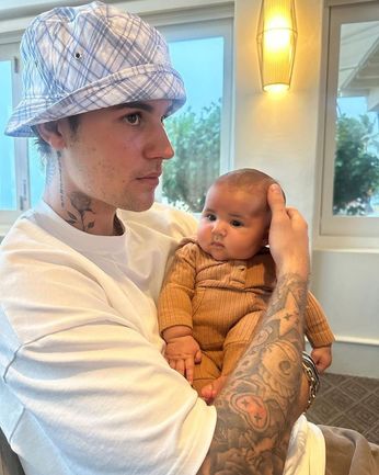 Justin Bieber in a white shirt and bucket hat, holding a baby in yellow, showing his tattoos indoors.
