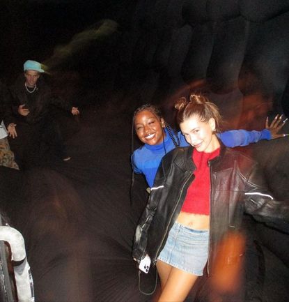 Two women posing with smiles, one with her arm around the other, in a dim, blurred setting with Justin Bieber in the background.