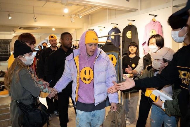 Justin Bieber in a yellow beanie and purple jacket shakes hands with a masked woman in a smiley-logo clothing store, surrounded by others.