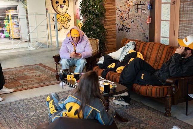 Three individuals in a graffiti-adorned room, with the possible presence of Justin Bieber in a black and yellow outfit reclining on a couch, while another person in a purple hoodie and yellow cap sits on a chair, and someone in a blue and yellow jacket is partially seen. The space exudes an urban, chill atmosphere with casual furnishings and bear-themed wall art.
