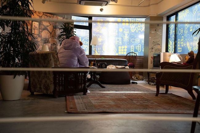 A cozy, eclectic room with Justin Bieber seated facing a brightly patterned window.