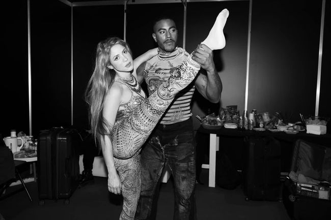Black and white photo of Shakira in body paint, backstage with a man who holds her leg as she leans back.