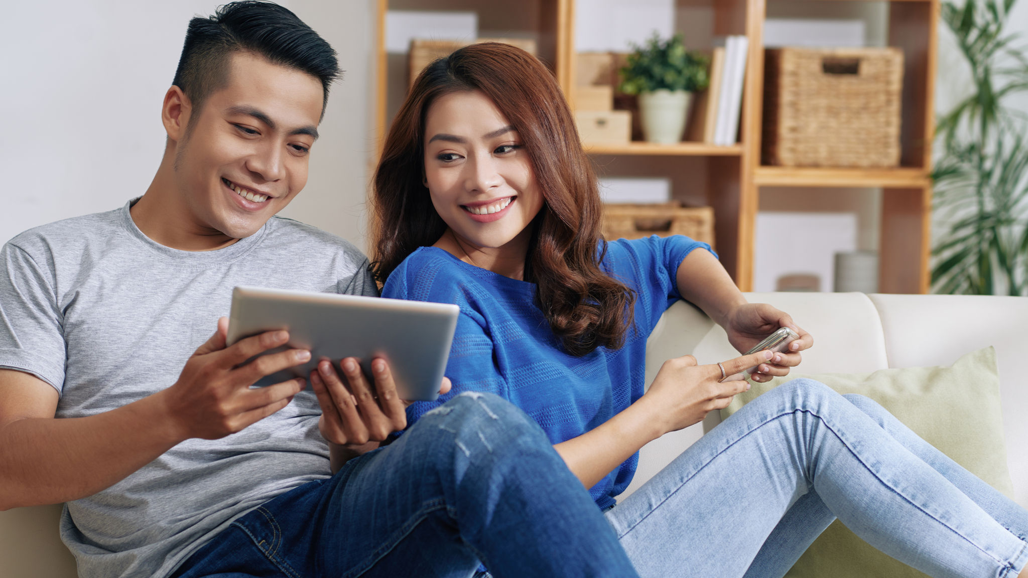 Smiling young man sitting on sofa and showing video on tablet to Asian girlfriend in living room