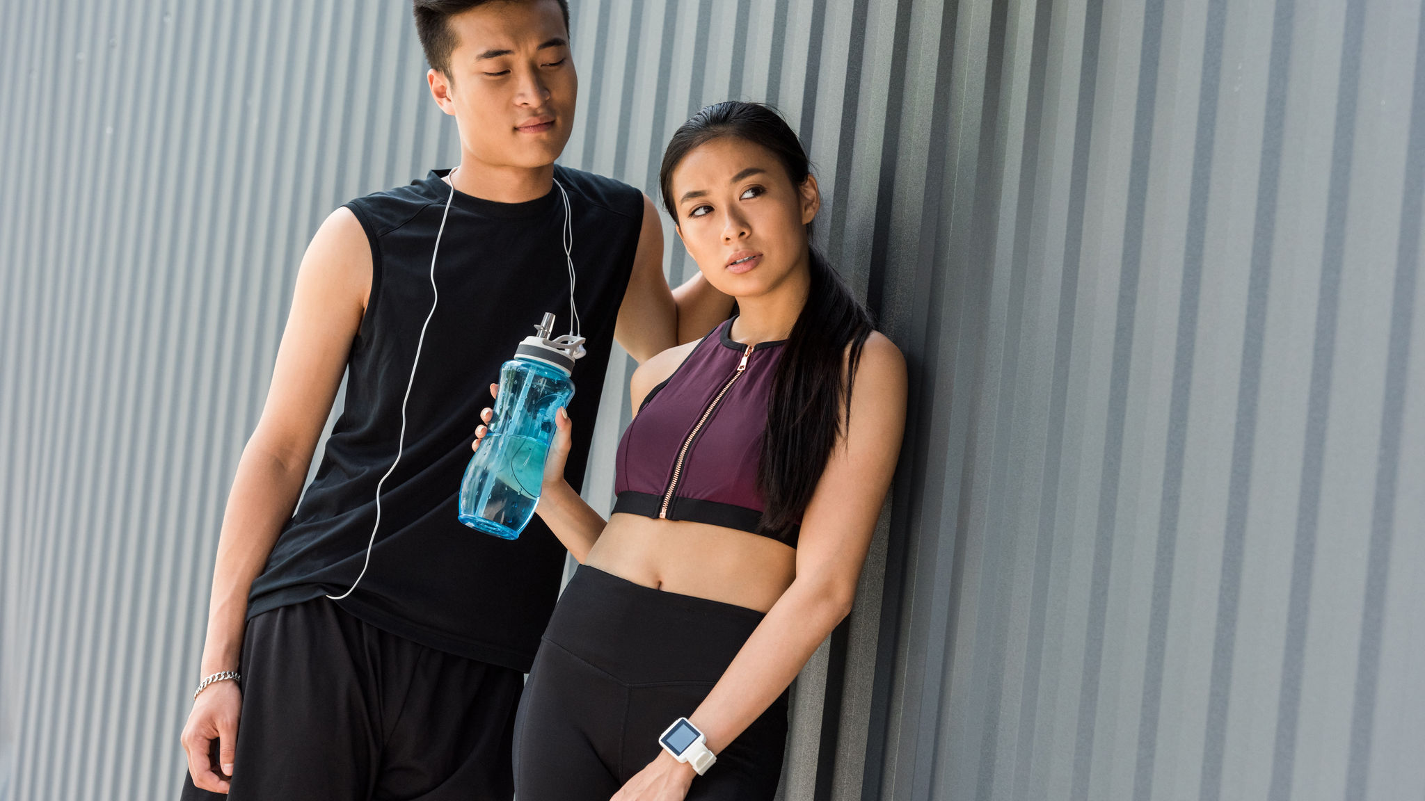 Smiling young Asian sportsman with earphones over neck standing near sportswoman with sport bottle