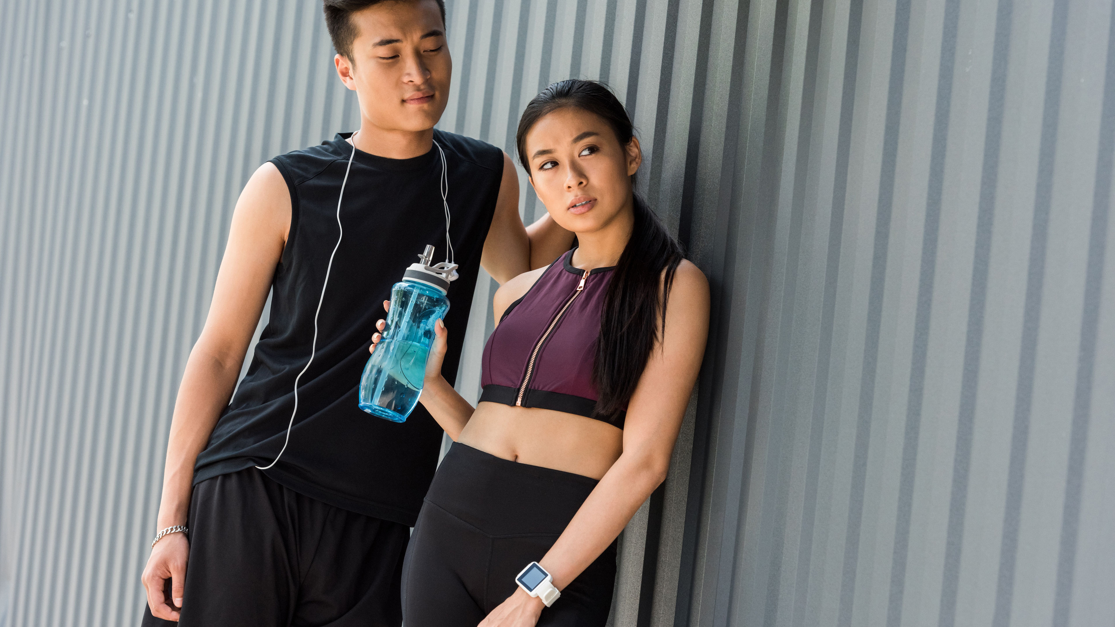 Smiling young Asian sportsman with earphones over neck standing near sportswoman with sport bottle